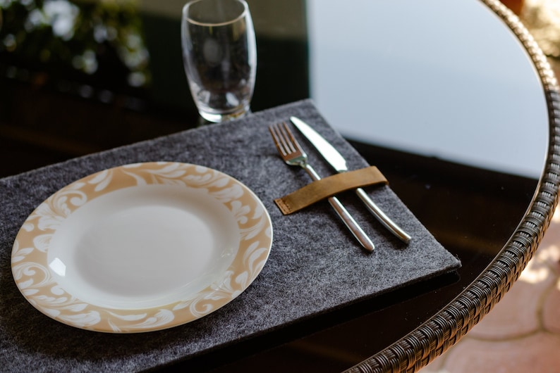 May include: A gray felt placemat with a white plate with a beige floral design. A silver fork and knife are placed on the placemat and held together with a brown leather strap.