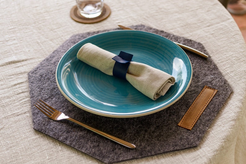 May include: A blue ceramic dinner plate with a white rim sits on a gray felt placemat. A white linen napkin is rolled and tied with a blue ribbon. A silver fork and a silver knife are on the placemat.