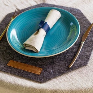 May include: A gray felt placemat with a leather tag, a blue plate with a white napkin, a silver fork, and a silver knife.