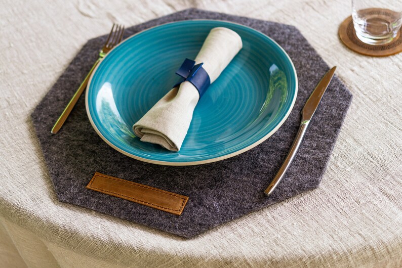 May include: A gray felt placemat with a leather tag, a blue plate with a white napkin, a silver fork, and a silver knife.