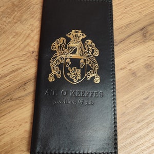 May include: Black leather card holder with gold embossed lettering and a crest. The text reads "A.T. O'Keeffe's Provisions & Pub".