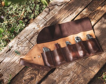 May include: A brown leather tool roll, partially unrolled, displaying various tools. The roll is made of a lighter tan leather on the inside and a darker brown on the outside. The tools include knives and other metal implements.