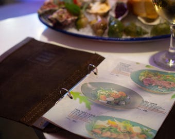 May include: A leather-bound menu is open, displaying colorful food images. A plate of oysters and a glass of white wine are in the background. The menu features images of salads and other dishes, suggesting a restaurant setting.