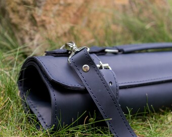 May include: A dark blue leather roll-up case with a strap and buckle closure. The case is lying on a bed of green grass.