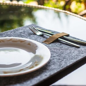 May include: A white ceramic plate with a gold floral design sits on a gray felt placemat. A leather strap holds a silver fork and knife together.