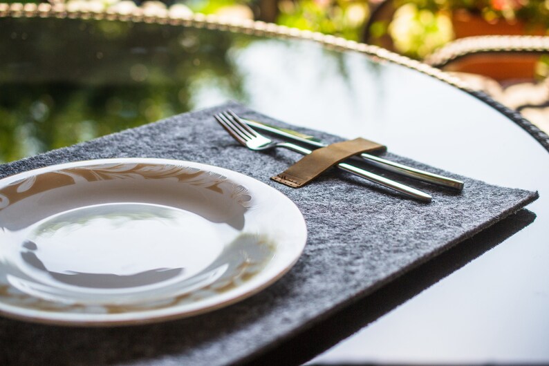 May include: A white ceramic plate with a gold floral design sits on a gray felt placemat. A leather strap holds a silver fork and knife together.