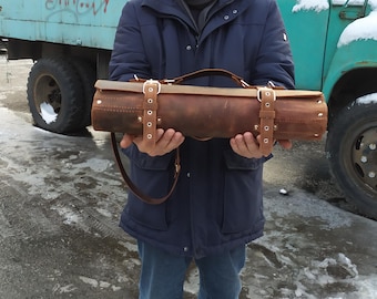 May include: A brown leather roll-up case with a strap and buckles. The case is being held by a person.
