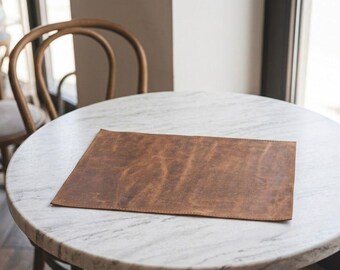 May include: A rectangular brown leather placemat with a distressed, mottled surface texture and visible stitched edges. The placemat features a natural grain pattern with variations in color ranging from light tan to deep chocolate brown tones.