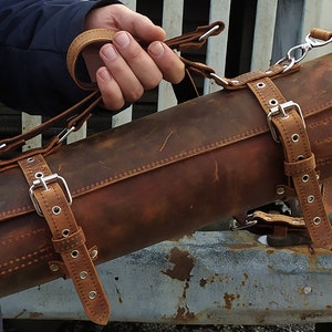 May include: A brown leather roll with multiple straps and buckles. The roll has a distressed look and is made of thick leather. The roll is being held by a hand.