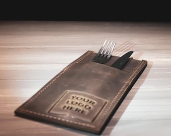 May include: A brown leather cutlery holder with a stitched edge. The holder has a pocket for a knife and fork and a space for a logo. The text "YOUR LOGO HERE" is printed on the holder.