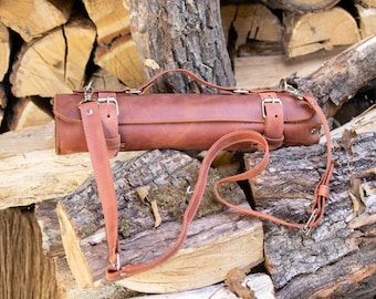 May include: A brown leather knife roll with a strap and buckles. The roll is sitting on a pile of logs.