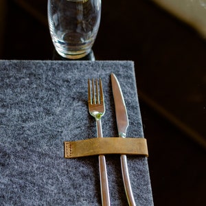 May include: A brown leather napkin ring holding a silver fork and knife on a gray felt placemat.