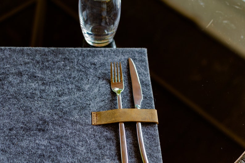 May include: A brown leather napkin ring holding a silver fork and knife on a gray felt placemat.
