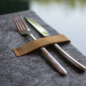 May include: A brown leather cutlery holder with a stitched edge, holding a silver fork and knife on a gray felt surface.