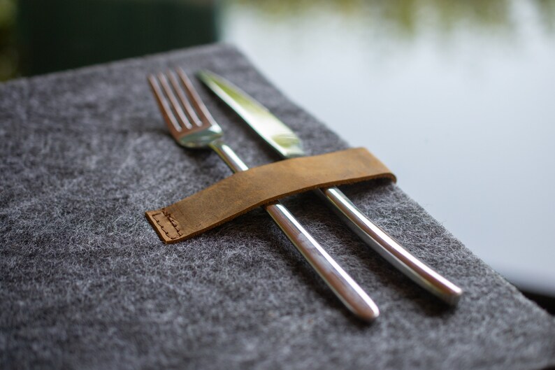 May include: A brown leather cutlery holder with a stitched edge, holding a silver fork and knife on a gray felt surface.