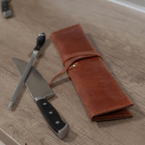 May include: A brown leather knife roll with a silver knife and a steel sharpening steel on a wooden surface.