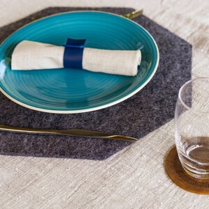 May include: A gray felt placemat with a blue plate, a white napkin, and a silver knife. A glass sits on a wooden coaster with a geometric design.