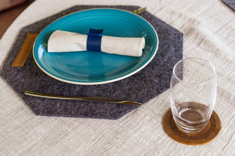 May include: A gray felt placemat with a blue plate, a white napkin, and a silver knife. A glass sits on a wooden coaster with a geometric design.