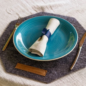 May include: A blue ceramic dinner plate with a white linen napkin tied with a blue ribbon sits on a gray felt placemat with a brown leather accent. A silver fork and knife are on the placemat.