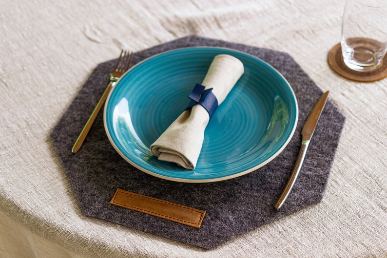 May include: A blue ceramic dinner plate with a white linen napkin tied with a blue ribbon sits on a gray felt placemat with a brown leather accent. A silver fork and knife are on the placemat.