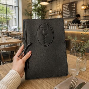 May include: A black leather menu cover with a textured surface and a central oval patch featuring an embossed corkscrew design. The rectangular cover is secured with a thin black cord tie on the left side, showcasing visible stitching along the edges.