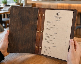 May include: A menu holder made of dark stained wood and brown leather, featuring brass screw posts. The menu page displays the text Oak & Leaf Kitchen, listing sections for Starters, Mains, and Desserts with various food items and prices.