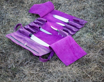 May include: A purple leather knife roll with several stainless steel knives. The roll is open, displaying the knives in individual slots. A matching purple towel is next to the roll. The roll has a zipper closure and a shoulder strap.