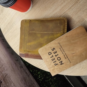 Puede incluir: Una cartera de cuero marrón con una pátina amarilla y una libreta marrón con el texto "FIELD NOTES" en la portada. La libreta está hecha en los EE. UU. y tiene 43 páginas.