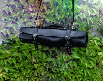 May include: A black leather knife roll with a strap and buckles. The roll is hanging from a hook.