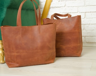 Könnte beinhalten: Zwei braune Ledertaschen mit langen Henkeln. Die Taschen stehen auf einem weißen Holzboden vor einer weißen Backsteinwand.