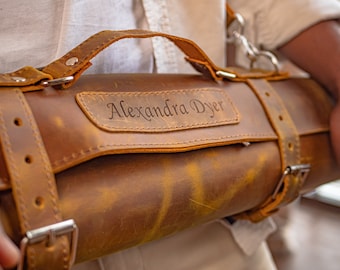 May include: A brown leather knife roll with a strap and buckle closure. The roll is personalized with the name "Alexandra Dyer" in a script font.