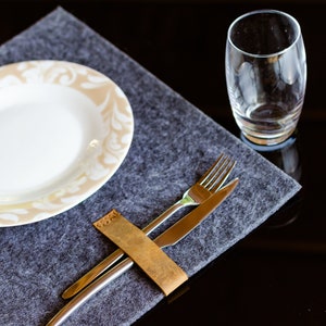 May include: A gray felt placemat with a white plate with a beige floral design. A leather wrapped fork and knife are placed on the placemat.