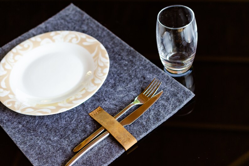 May include: A gray felt placemat with a white plate with a beige floral design. A leather wrapped fork and knife are placed on the placemat.