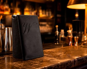 May include: A black, rectangular, upright menu holder with a matte finish and a stylized letter W embossed on the front cover. The item features stitched edges and a vertical orientation, standing on a surface with a dark, reflective texture.