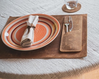 May include: A brown leather placemat with a stitched pocket for cutlery. A white napkin is rolled and placed on a red and white striped ceramic plate.