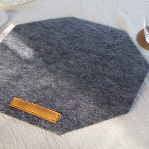 May include: A gray octagonal felt placemat with a brown leather label. The placemat is on a white linen tablecloth.