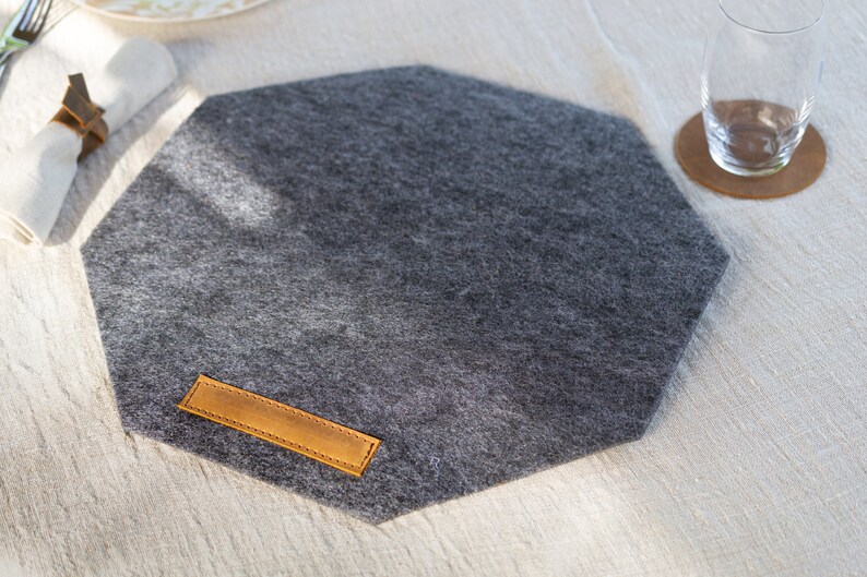 May include: A gray octagonal felt placemat with a brown leather label. The placemat is on a white linen tablecloth.