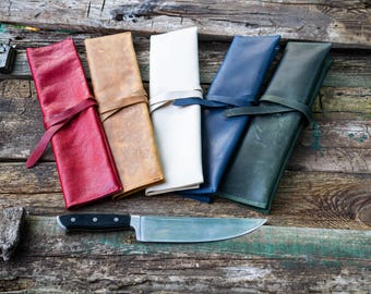 May include: Six leather knife rolls in different colors, including red, brown, white, blue, and green, laid out on a wooden surface. A silver knife is placed in front of the rolls.