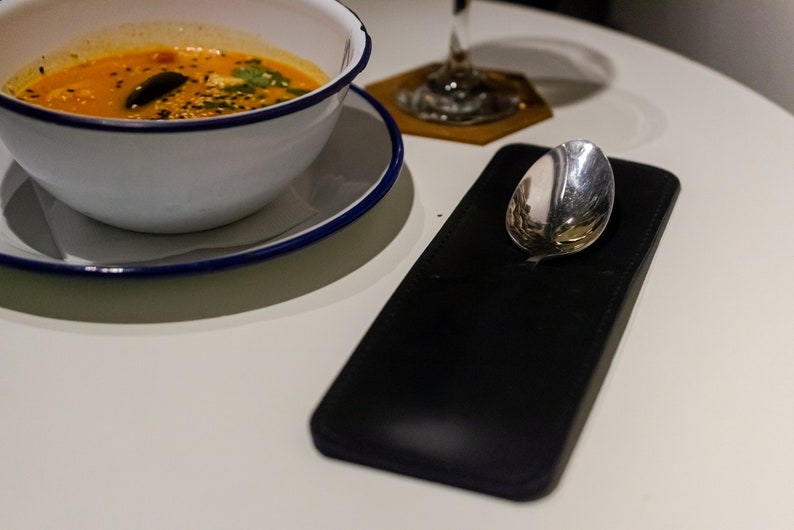 Pu&ograve; includere: Una ciotola bianca con un bordo blu piena di zuppa arancione e guarnita con semi di sesamo neri ed erbe verdi. Un portacucchiaio in pelle nera con un cucchiaio d'argento su un tavolo bianco.
