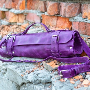 May include: A purple leather roll-up bag with multiple buckles and a long strap. The bag is lying on a gray concrete surface with broken red bricks in the background.