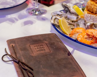 May include: A brown leather menu cover with a strap closure. The cover has the text "YOUR LOGO HERE" embossed on the front. The menu cover is on a white tablecloth next to a plate of oysters, shrimp, and lemon.