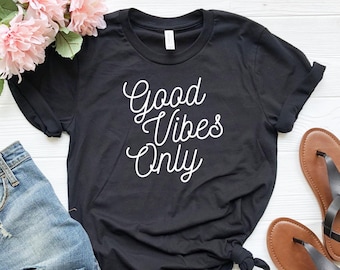 May include: Black t-shirt with white text that reads "Good Vibes Only". The shirt is laid flat on a wooden surface with a pair of blue jeans and sandals.