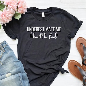 May include: Black t-shirt with white text that reads "UNDERESTIMATE ME (that'll be fun!)". The shirt is laid flat on a white surface with a pair of blue jeans and brown sandals.