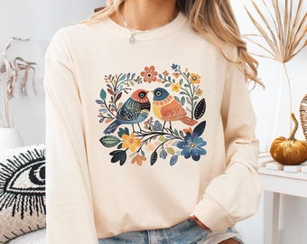 May include: A cream-colored long-sleeved shirt with a colorful floral and bird design. The birds are blue, red, and yellow, and the flowers are pink, yellow, and blue.