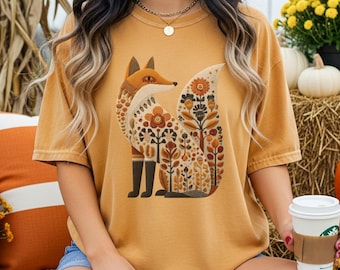 May include: Mustard-colored t-shirt featuring a stylized fox design with floral patterns in shades of brown, orange, and cream. The fox is wearing black boots. The shirt is a relaxed fit and has short sleeves. The word "Mustard" is printed at the bottom.