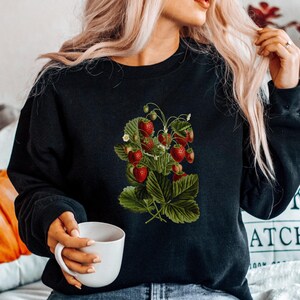 May include: A black sweatshirt with a vintage illustration of strawberries on the front. The sweatshirt is worn by a person with long blonde hair and blue jeans.