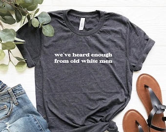 May include: Dark grey heather t-shirt with the text "we've heard enough from old white men" in white. The shirt is paired with blue jeans and brown sandals with black straps. The words "DARK GREY HEATHER" are printed at the bottom.