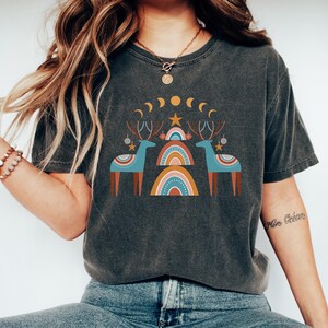 Puede incluir: Una camiseta gris oscuro con un dise&ntilde;o gr&aacute;fico que presenta dos renos estilizados de pie frente a un arco&iacute;ris. El dise&ntilde;o tambi&eacute;n incluye una estrella, lunas crecientes y peque&ntilde;os c&iacute;rculos.