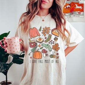 May include: A white t-shirt with a fall-themed design featuring illustrations of a pumpkin, leaves, a mug of hot chocolate, a sweater, a scarf, and a pie. The text "I love fall most of all" is printed on the shirt.