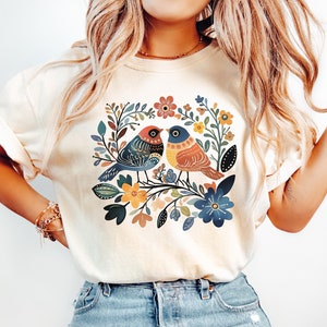 May include: Ivory-colored sweatshirt featuring a colorful floral and bird design. The graphic includes two birds perched on a branch surrounded by flowers in shades of blue, orange, and green. The word "ivory" is printed at the bottom.